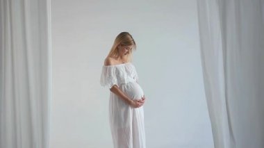 Genç, hamile, beyaz elbiseli bir anne, karnında bebeğine sarılıyor. Kolay, sağlıklı, uzun zamandır beklenen hamilelik kavramı. Anneliğin mutluluğu. Taşıyıcı anne.