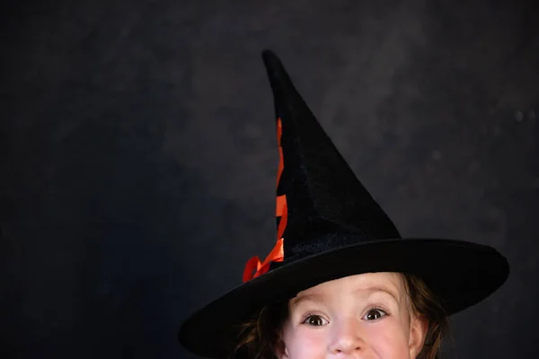 Close-up portrait of a Little happy girl in a black witch hat against the background of a dark concrete grunge wall in loft. Child\'s Halloween costume. Empty space for text. Mock up and copy space.