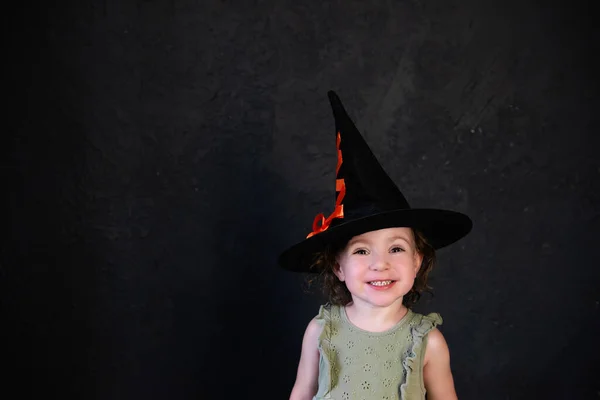 一个戴着黑色女巫帽的快乐小女孩的特写镜头 背景是阁楼里黑暗的混凝土墙体 孩子的万圣节服装 空白的文字空间 模拟和复制空间 — 图库照片