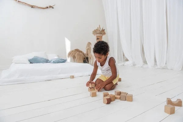 Little happy boy Black African American play houses with wooden eco bricks, build and destroy towers. Fine motor development, educational games. White interior in background with wooden floor