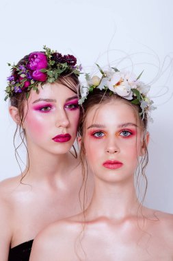 Parlak ve moda bahar sanatları iki güzel kız kardeş için profesyonel makyaj. Portreyi kapat. Taze bahar çiçeklerinden oluşan bir halka kafalarda şakayık. Temiz ciltli, yüz bakımlı bir model. Gençler için kozmetoloji