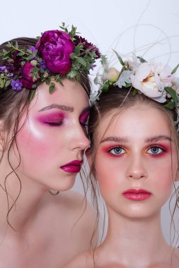 Parlak ve moda bahar sanatları iki güzel kız kardeş için profesyonel makyaj. Portreyi kapat. Taze bahar çiçeklerinden oluşan bir halka kafalarda şakayık. Temiz ciltli, yüz bakımlı bir model. Gençler için kozmetoloji