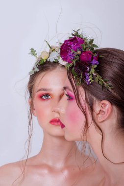 Parlak ve moda bahar sanatları iki güzel kız kardeş için profesyonel makyaj. Portreyi kapat. Taze bahar çiçeklerinden oluşan bir halka kafalarda şakayık. Temiz ciltli, yüz bakımlı bir model. Gençler için kozmetoloji
