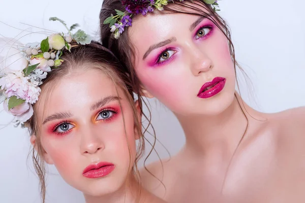 Parlak ve moda bahar sanatları iki güzel kız kardeş için profesyonel makyaj. Portreyi kapat. Taze bahar çiçeklerinden oluşan bir halka kafalarda şakayık. Temiz ciltli, yüz bakımlı bir model. Gençler için kozmetoloji