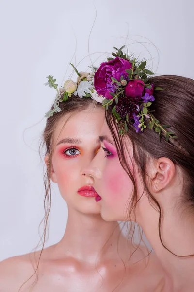 Parlak ve moda bahar sanatları iki güzel kız kardeş için profesyonel makyaj. Portreyi kapat. Taze bahar çiçeklerinden oluşan bir halka kafalarda şakayık. Temiz ciltli, yüz bakımlı bir model. Gençler için kozmetoloji