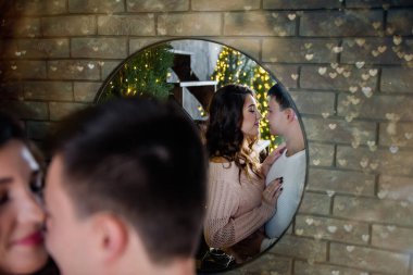 Yuvarlak bir aynada, öpüşen romantik bir çiftin yakın plan portresinin yansıması. Tuğla duvarın arka planına sarılan bir çift. Işıklardan bir çelenk, Noel ağaçları. Uzayı kopyala Yeni Yılda Öpücük 