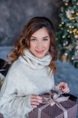 Kıvırcık kahverengi saçlı, Noel ağacı ve şöminenin yanında oturan, elinde bir hediye tutan modern bir genç kadın. Etrafta bir sürü hediye kutusu var. Arka planda çelenkler parlıyor. Yeni yıl arifesi. Kart