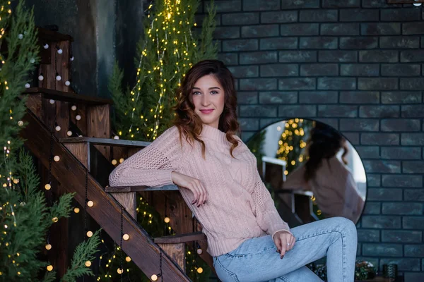 柔らかいピンクのニットセーターを着た女の子 巻き毛で長い髪の青いジーンズは クリスマスのモミの木や照明付きのガーランドの間で木製の階段に座って 広く笑顔 若い女性のクローズアップポートレート — ストック写真