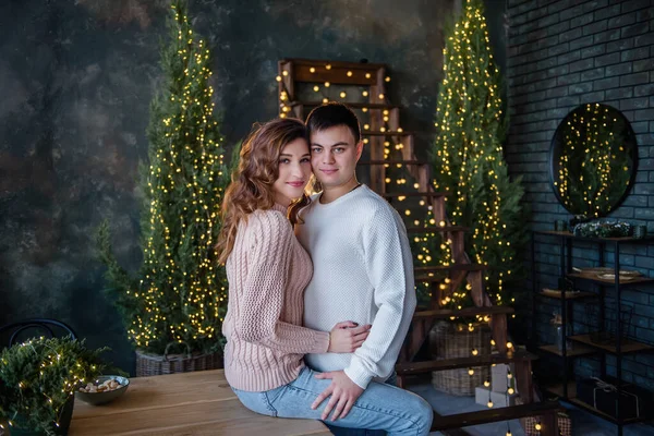 Pembe örgü süveterli bir kız ahşap bir masada oturuyor, beyazlar içindeki genç bir adam ona sarılıyor. Yeni yıl arifesinde sevgi dolu bir çift kucaklaşması, öpücükler. Işık çelenkleri, Noel ağaçları