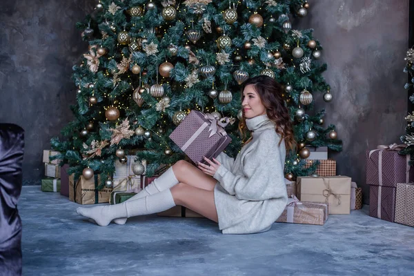 巻き茶色の髪をした現代の若い女性は クリスマスツリーと暖炉のそばに座って プレゼントを持っています ギフトボックスはたくさんあります 背景にはガーランドが輝いています 大晦日だよ カード — ストック写真