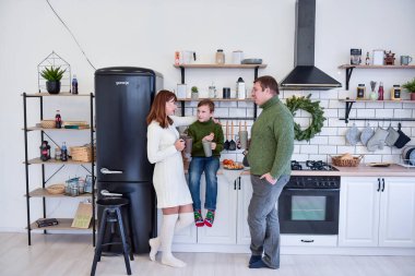 Beyaz mutfakta İskandinav tarzında mutlu bir aile, sıcak içecekler içip gülüyorlar. Genç bir kadın oğluna sarılıyor, bir adam elinde gri bir bardak tutuyor. Minimalist Noel çelengi. Boşluğu kopyala 