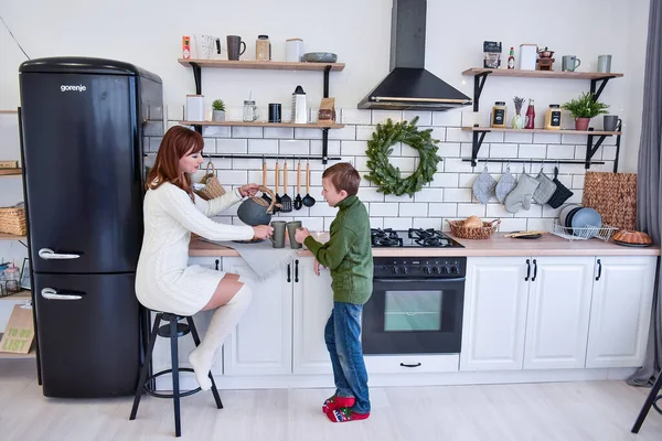 A young woman is sitting in a modern kitchen with her son. The family drinks a hot drink tea coffee, laughs. Minimalistic scandinavian style, black refrigerator, green christmas wreath on brick wall