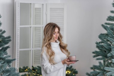 Sıcak kış süveteri giymiş mutlu bir kız İskandinav tarzı bir mutfakta duruyor ve Noel ağacının yanındaki pembe bardaktan sıcak bir içecek içiyor. Yeni yıl sabahı. Şenlikli iç ve ev dekorasyonu.
