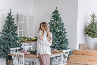 Güzel kıvırcık saçlı, uzun saçlı, Noel ağaçlarının yanında ışık çelenkleriyle. Gri örgü süveter giymiş, büyük yakalı, pudralı elbisesi olan neşeli bir kız mutfakta oturuyor.