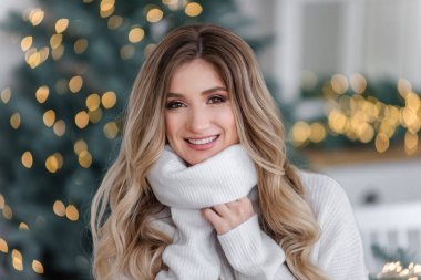 Güzel kıvırcık saçlı, uzun saçlı, Noel ağaçlarının yanında ışık çelenkleriyle. Gri örgü süveter ve büyük yakalı beyaz gülüşlü bir kızın yakın plan portresi. Boşluğu, dokuyu kopyala