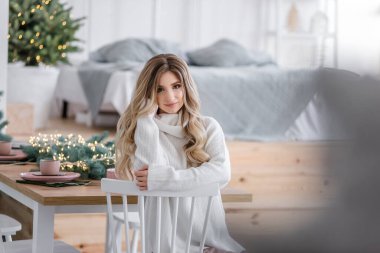 Güzel kıvırcık saçlı, uzun saçlı, Noel ağaçlarının yanında ışık çelenkleriyle. Gri örgü süveter ve büyük yakalı beyaz gülüşlü bir kızın yakın plan portresi. Boşluğu, dokuyu kopyala