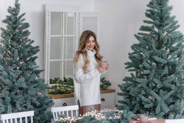 暖かい冬のセーターを着た幸せな女の子が北欧風のキッチンに立って クリスマスツリーのピンクのカップから熱い飲み物を飲みます お正月の朝 祭りのインテリアと家の装飾 — ストック写真