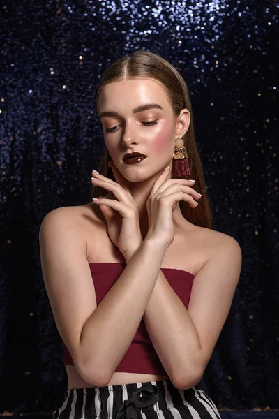 Fashionable girl with bright, professional evening make-up. Burgundy lipstick, clean, natural face, French manicure. Hair is parted, large hanging gold earrings with pearls. Shiny  blue background