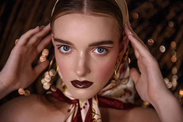 Close-up portrait of a girl with evening, fashion make-up, maroon lipstick, clear skin, pink rumanian, round gold earrings in her ears, head covered with a fashionable satin headscarf. Golden glitter