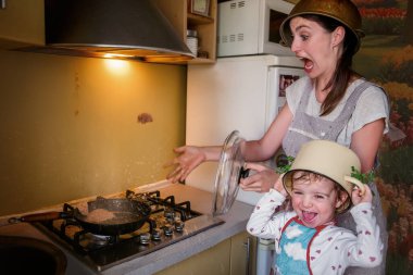 Anne ve kızı mutfakta deli gibi pirzola kızartıyorlar. Çocuklarla yemek pişirmek eğlenceliydi. Yağ her yöne sıçrar. Kirli mutfak paneli ve duvar. Kızların kafalarında tavalar var. Mizah kavramı