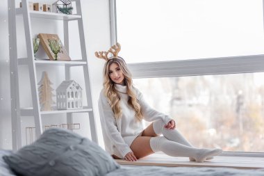 Sarı Noel geyiği boynuzları olan, gri örgü kazaklı, uzun taytlı sarışın bir kız panoramik pencerenin yanındaki pencere eşiğinde oturuyor. Ev heykelcikleri olan beyaz bir askı var. Uzayı kopyala bulanık