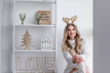 Sarı Noel geyiği boynuzları olan, gri örgü kazaklı, uzun taytlı sarışın bir kız panoramik pencerenin yanındaki pencere eşiğinde oturuyor. Ev heykelcikleri olan beyaz bir askı var. Uzayı kopyala bulanık