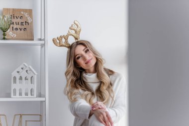 Sarı Noel geyiği boynuzları olan, gri örgü kazaklı, uzun taytlı sarışın bir kız panoramik pencerenin yanındaki pencere eşiğinde oturuyor. Ev heykelcikleri olan beyaz bir askı var. Uzayı kopyala bulanık