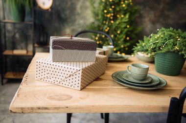 Tahta bir eko masanın üzerinde güzel bir paketin içinde fiyonklu hediyeler var. Yakınlarda küçük Noel ağaçları olan yeşil bir seramik tabak, ışıktan çelenkleri olan bir thuja var. Boşluğu, kartı, şablonu kopyala