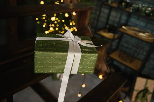 盒子用绿色礼品纸包裹着 装饰有彩带和米色的蝴蝶结 圣诞包装的背景是模糊的假牙与灯 复制空间 — 图库照片