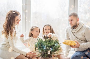 Beyaz kazaklı mutlu bir aile panoramik pencereli güzel bir çatı katında yeni yıla hazırlanıyor. Küçük kızları olan genç ebeveynler küçük bir Noel ağacını tencerede süslüyorlar. Uzayı kopyala, bokeh