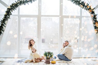Beyaz kazaklı mutlu bir aile panoramik pencereli güzel bir çatı katında yeni yıla hazırlanıyor. Küçük kızları olan genç ebeveynler küçük bir Noel ağacını tencerede süslüyorlar. Uzayı kopyala, bokeh