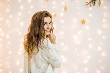 Uzun kıvırcık saçlı güzel bir kadın, parlak ışık çelenkleri ve bir Noel ağacının dalları karşısında mutlu bir şekilde gülümsüyor. Kız beyaz örgü süveter ve kot pantolon giyiyor.