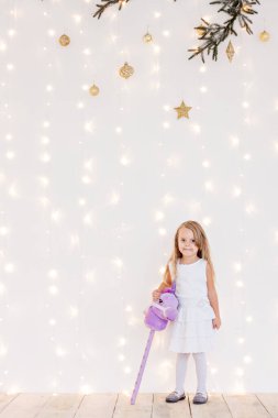Beyaz bir şenlik elbisesi içindeki küçük sarışın kız Noel ağacının ışıklarından sarı bokeh bir arka planda. Yeni yıl kartı. Bebek gülümsüyor, eğleniyor, elinde mor oyuncak at bastonu tutuyor. Boşluğu kopyala. 