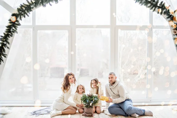 Beyaz kazaklı mutlu bir aile panoramik pencereli güzel bir çatı katında yeni yıla hazırlanıyor. Küçük kızları olan genç ebeveynler küçük bir Noel ağacını tencerede süslüyorlar. Uzayı kopyala, bokeh