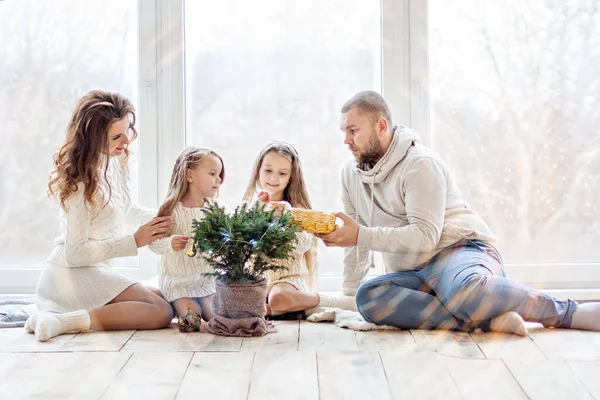 Beyaz kazaklı mutlu bir aile panoramik pencereli güzel bir çatı katında yeni yıla hazırlanıyor. Küçük kızları olan genç ebeveynler küçük bir Noel ağacını tencerede süslüyorlar. Uzayı kopyala, bokeh