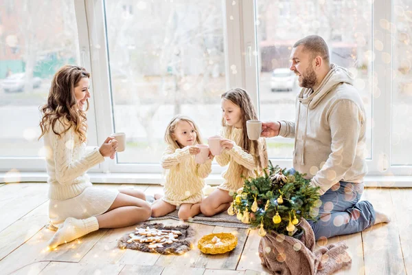 Happy family in white sweaters drinks cocoa by the panoramic window. Parents and daughters are holding cups in their hands, on a plate are gingerbread men near a small Christmas tree in pot. New year