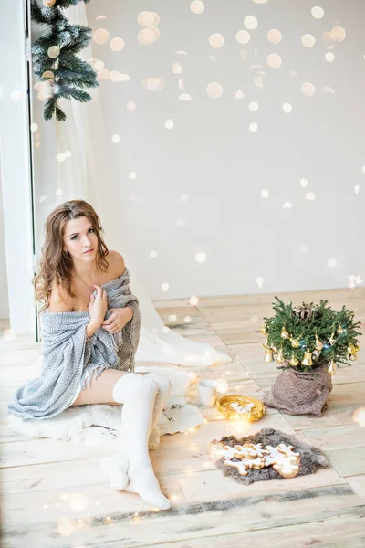 官能的な若い女性が灰色の再生に包まれた自分自身を保持している クリスマスツリー ガーランドの枝で飾られたパノラマの窓に座っています 少女は白いレギンスで裸になっている 冬休み — ストック写真