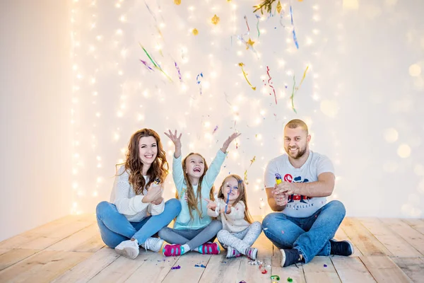 Mutlu aile, sarı ışık çelenkleri ve bokeh Noel ağacı dallarının arka planında oynaşarak eğleniyor. İyi yeni yıl ruhu. Ebeveynlerin kızları konfeti atıyor, sarılıyor, gülüyor. Boşluğu kopyala
