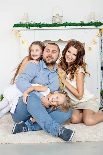 A happy family is sitting on the carpet by the white fireplace with an elegant Christmas tree in garlands of lights. Young father and beautiful mother hug little sisters, play, have fun. Copy space