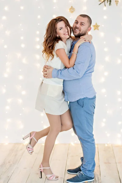 A loving couple have fun on the background of a white wall with garlands of bright lights, a branch of a Christmas tree hangs from above. Lovers are happy dancing at the party waiting for the new year