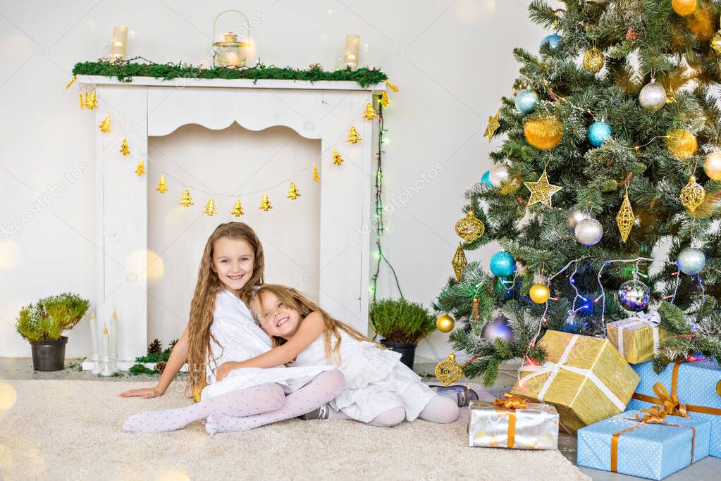 Two little blonde girls in white dresses are playing by the fireplace and Christmas tree with boxes. Sisters have fun, hug, laugh, unfold gifts from Santa Claus. New year card. Copy space. Friendship