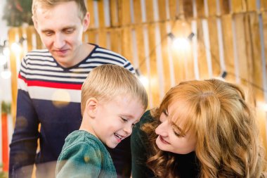 Mutlu bir aile, Noel dalları, çelenkler ve çam iğneleri ile süslenmiş kırmızı bir arabanın yanında oğluyla eğleniyor ve oynuyor. Anne ve baba çocuğa sarılıyor. Yeni Yıl