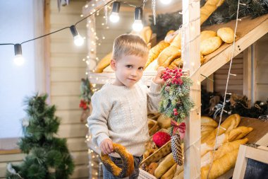 Örgü beyaz süveter giyen küçük bir çocuk fırında merdiven üzerinde duruyor, hamur işlerini seçiyor, gülüyor. Gösteri Noel ağaçları, yeni yıl çelenkleri, kar ve buz saçaklarıyla süslendi. Taze ekmek. 