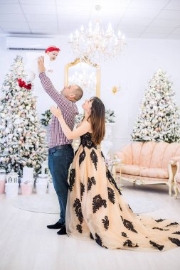 Genç bir aile akşam için şen şakrak giyiniyor, kucağında küçük bir bebek var. Arkaplandaki karla kaplı Noel ağaçlarına ışık çelenkleri, hediyeler, beyaz bir şömine. Kart