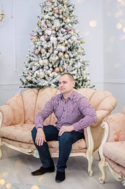 Orta yaşlı, ekose gömlekli genç bir adam, modern bir koltukta, Noel ağacının arka planında, ışıklardan oluşan beyaz bir şömine, altın oyuncaklar ve top ile oturuyor. Mutlu, gülümseyen. Boşluğu kopyala