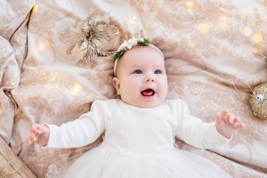 Kafasında süs olan beyaz elbiseli küçük bir bebeğin yakın plan portresi. Kız ışıklardan oluşan çelenklerle bir kanepede yatıyor, neşeli bir Noel ağacı, gülüyor. Boşluğu kopyala, tebrik kartı
