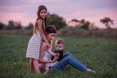 Anne oğlunu ve kızını kucaklıyor, alacakaranlık çökerken çimen tarlasında oturuyor. Huzurlu bir sevgi, bir bağ anı. Dusk 'ta Serene Family Time. Yeşil çayırda çocuklara sarılan bir kadın. Anneler Günü