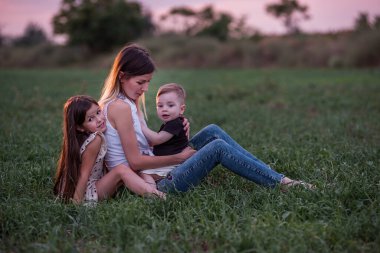 Anne oğlunu ve kızını kucaklıyor, alacakaranlık çökerken çimen tarlasında oturuyor. Huzurlu bir sevgi, bir bağ anı. Dusk 'ta Serene Family Time. Yeşil çayırda çocuklara sarılan bir kadın. Anneler Günü
