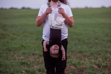 Farklılık babası küçük oğlunu baş aşağı tutarken neşeli çocuğunu gösteriyor. Uzun kıvırcık saçlı, sakallı, gözlüklü gün batımı yeşil tarlasında bebekle oynaşan genç bir adam.