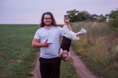 Farklılık babası küçük oğlunu baş aşağı tutarken neşeli çocuğunu gösteriyor. Uzun kıvırcık saçlı, sakallı, gözlüklü gün batımı yeşil tarlasında bebekle oynaşan genç bir adam.